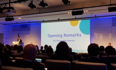 A screen which reads 'Opening remarks', with Pia Ahrenkilde Hansen standing at a lectern to the left