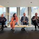 Six people sit on a stage, João Costa is holding a microphone and speaking 