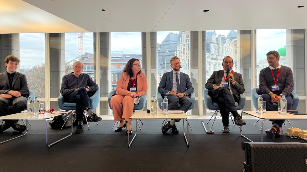 Six people sit on a stage, João Costa is holding a microphone and speaking 