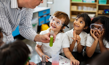 Three children wearing safety goggles smile at their teacher who is holding a test tube with a green liquid inside